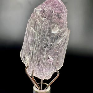 Puede incluir: Un esp&eacute;cimen de cristal transl&uacute;cido, de color p&uacute;rpura claro, montado sobre un soporte met&aacute;lico plateado. El cristal tiene una superficie rugosa y texturizada con estructuras internas visibles. El fondo es gris oscuro, creando contraste.