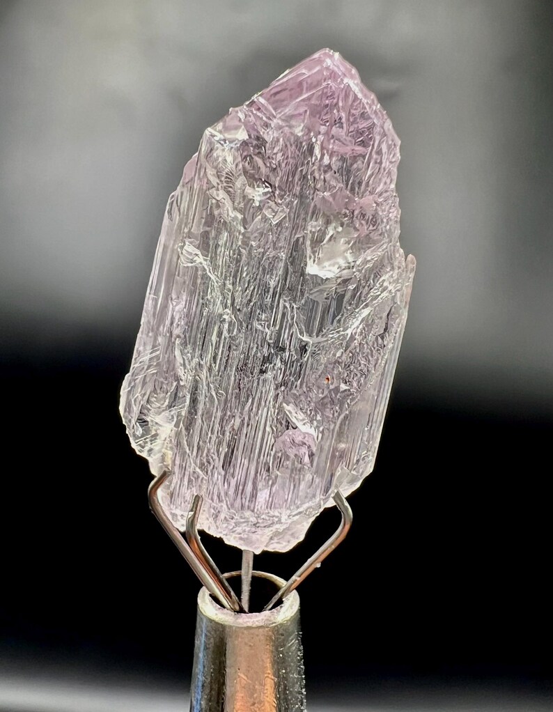 Puede incluir: Un esp&eacute;cimen de cristal transl&uacute;cido, de color p&uacute;rpura claro, montado sobre un soporte met&aacute;lico plateado. El cristal tiene una superficie rugosa y texturizada con estructuras internas visibles. El fondo es gris oscuro, creando contraste.