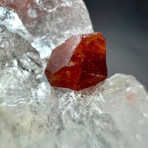 Könnte beinhalten: Nahaufnahme einer rohen, orange-roten Kristallformation. Der Kristall hat eine tiefe, satte Farbe und eine facettierte, geometrische Form. Er liegt auf einem durchscheinenden, eisig aussehenden Mineral, was einen auffälligen Kontrast erzeugt.