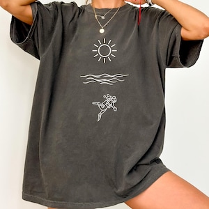 May include: Dark gray t-shirt with a white graphic of a sun, waves, and a scuba diver. The shirt has short sleeves and a relaxed fit. The design is simple and minimalist, perfect for casual wear.