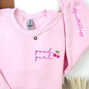 Puede incluir: Una sudadera rosa claro con las palabras "good girl" y un gr&aacute;fico de cerezas bordado en rosa en la parte delantera. La manga derecha tiene la palabra "Sparklellagg" bordada en rosa. Un collar dorado est&aacute; drapeado sobre la sudadera.
