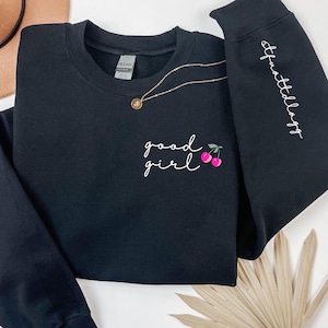 Puede incluir: Sudadera negra con las palabras "good girl" en escritura blanca, acompa&ntilde;ada de cerezas rosas. La manga derecha tiene el texto "#thatldaygg" en escritura blanca. Un collar dorado con un colgante est&aacute; sobre la sudadera.