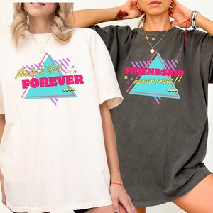 May include: Two t-shirts, one cream and one charcoal, each featuring a retro design. The cream shirt displays "Make it last FOREVER" in a pink, teal, and yellow triangle. The charcoal shirt says "FRIENDSHIP never ends!!" in a similar style.