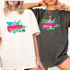 May include: Two t-shirts, one cream and one charcoal, feature a retro graphic. The cream shirt says "Drunk in Love" with a roller skate and other icons. The charcoal shirt says "Just Drunk" with a boombox and other icons. Both shirts have a vintage style.