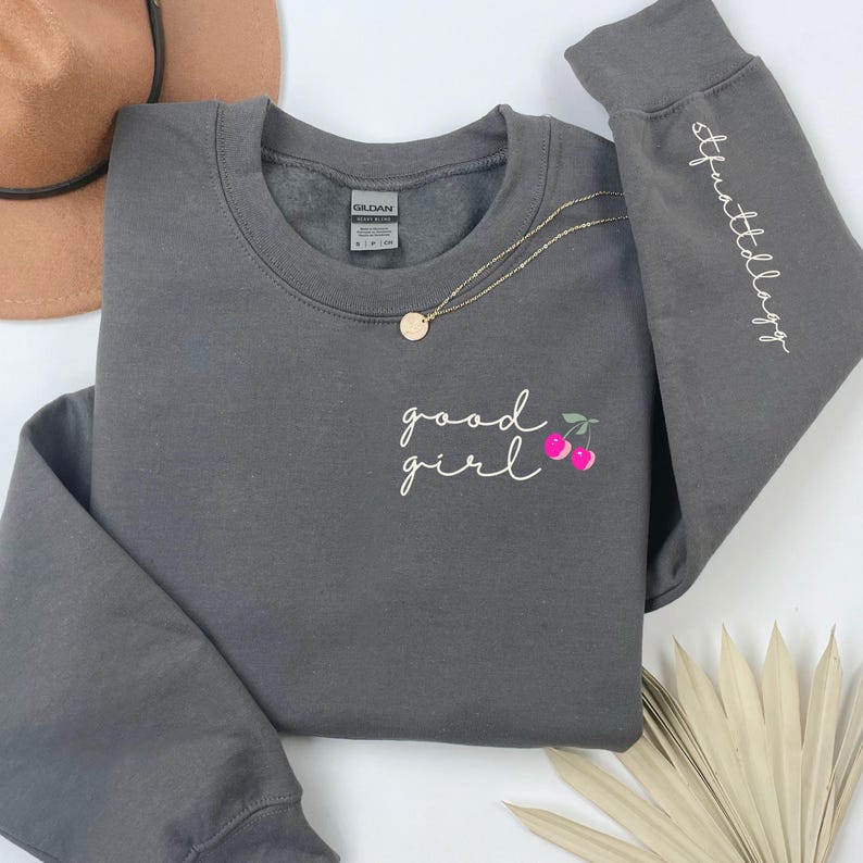 Puede incluir: Una sudadera gris carb&oacute;n con las palabras "good girl" en escritura blanca y un gr&aacute;fico de cerezas. La manga tiene el texto "stfuttldogg" en escritura blanca. Tambi&eacute;n se ven un collar dorado y un sombrero marr&oacute;n.