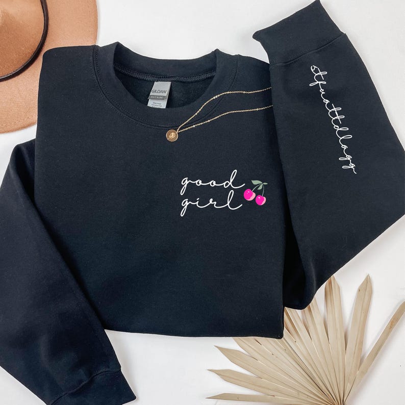 Puede incluir: Sudadera negra con las palabras "good girl" en escritura blanca, acompa&ntilde;ada de cerezas rosas. La manga derecha tiene el texto "#thatldaygg" en escritura blanca. Un collar dorado con un colgante est&aacute; sobre la sudadera.
