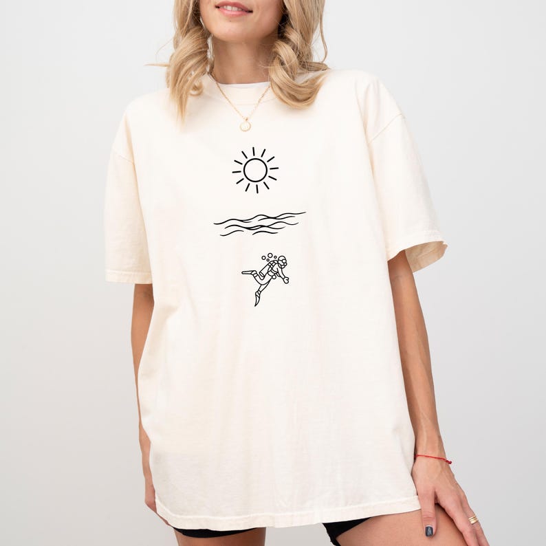 May include: Cream-colored t-shirt featuring a black line drawing of a sun, waves, and a scuba diver. The shirt has short sleeves and a relaxed fit. The design is simple and minimalist, suitable for casual wear.