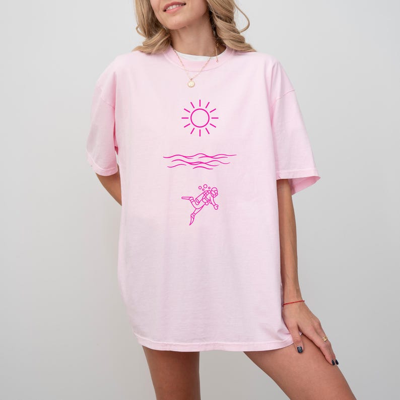 May include: A light pink t-shirt featuring a magenta graphic of a sun, waves, and a scuba diver. The design is a simple line drawing. The shirt is a relaxed fit.