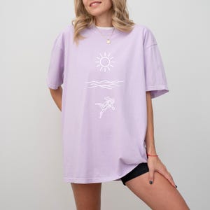May include: A light purple t-shirt with a white graphic of a sun, waves, and a scuba diver. The shirt has short sleeves and a relaxed fit.