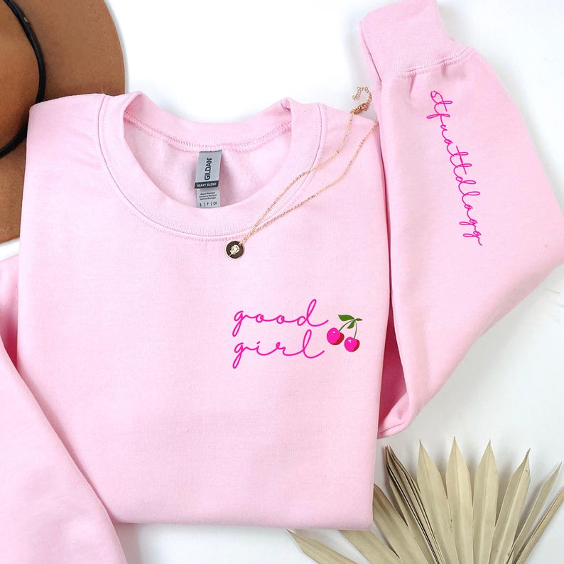 Puede incluir: Una sudadera rosa claro con las palabras "good girl" y un gr&aacute;fico de cerezas bordado en rosa en la parte delantera. La manga derecha tiene la palabra "Sparklellagg" bordada en rosa. Un collar dorado est&aacute; drapeado sobre la sudadera.
