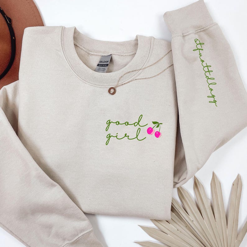 Puede incluir: Una sudadera beige con las palabras "good girl" en escritura verde, acompa&ntilde;ada de cerezas rosas. La manga derecha presenta la palabra "stuffattlaggs" en escritura verde. Un collar dorado con un colgante est&aacute; sobre la sudadera.