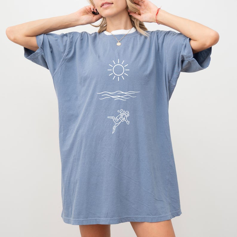 May include: A blue t-shirt with a white graphic of a sun, waves, and a scuba diver. The shirt has a white collar and short sleeves. The model is wearing a necklace and a red bracelet.