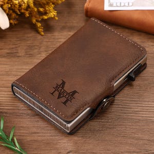 Puede incluir: Cartera de cuero marrón con el nombre "MARTIN" y la letra "M" grabados en la parte delantera. La cartera tiene un marco de metal y un cierre de botón. La cartera está sobre una superficie de madera.