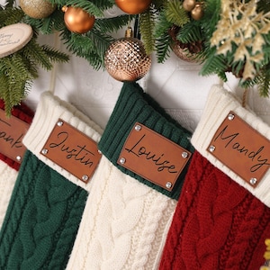 Puede incluir: Cuatro calcetines navideños de punto trenzado en crema, verde y rojo, cada uno con una etiqueta de nombre de cuero personalizada. Los calcetines cuelgan de una guirnalda con adornos y vegetación, creando una exhibición festiva.