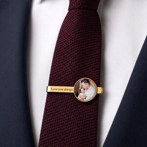 May include: A gold-colored tie clip with a circular photo and the words "Love you always." The tie clip is on a burgundy tie, set against a white shirt and a navy blue suit jacket.