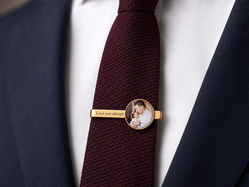 May include: A gold-colored tie clip with a circular photo and the words "Love you always." The tie clip is on a burgundy tie, set against a white shirt and a navy blue suit jacket.