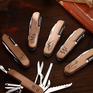 Puede incluir: Una colecci&oacute;n de navajas de bolsillo de madera personalizadas con varios grabados, incluyendo iniciales, nombres y un dise&ntilde;o de monta&ntilde;a. Las navajas est&aacute;n dispuestas sobre una superficie de madera oscura, con un libro y una botella en el fondo.