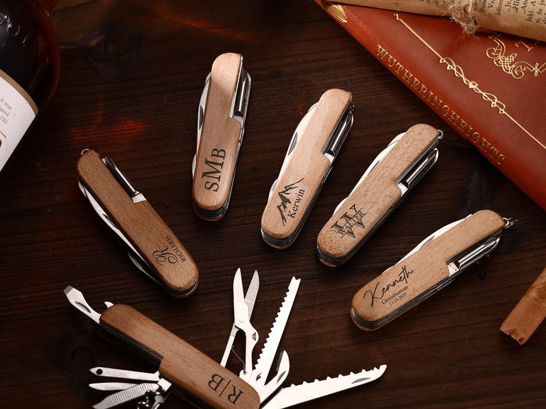 Puede incluir: Una colecci&oacute;n de navajas de bolsillo de madera personalizadas con varios grabados, incluyendo iniciales, nombres y un dise&ntilde;o de monta&ntilde;a. Las navajas est&aacute;n dispuestas sobre una superficie de madera oscura, con un libro y una botella en el fondo.