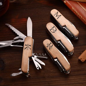 Puede incluir: Una colecci&oacute;n de navajas de bolsillo de madera personalizadas con varias herramientas, incluyendo una hoja de cuchillo, sierra y sacacorchos. Cada navaja presenta una inicial diferente grabada en el mango de madera. Las navajas est&aacute;n sobre una superficie de madera oscura.