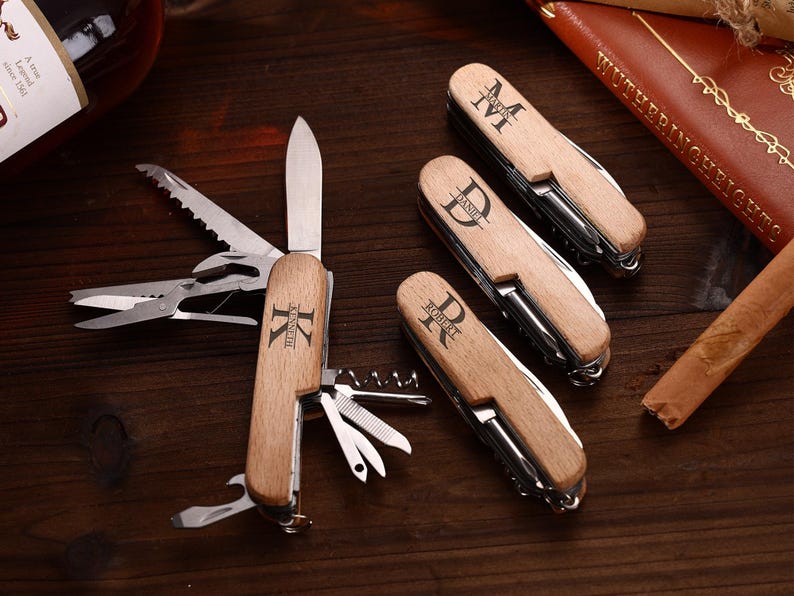 Puede incluir: Una colecci&oacute;n de navajas de bolsillo de madera personalizadas con varias herramientas, incluyendo una hoja de cuchillo, sierra y sacacorchos. Cada navaja presenta una inicial diferente grabada en el mango de madera. Las navajas est&aacute;n sobre una superficie de madera oscura.