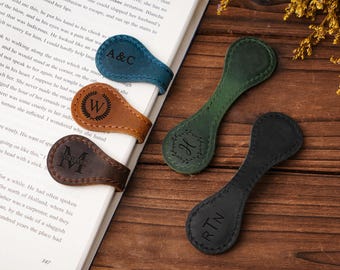 Puede incluir: Marcadores magnéticos de cuero en verde azulado, marrón, marrón oscuro, verde y negro. Cada marcador presenta una inicial o monograma único. Los marcadores están sobre un libro y una superficie de madera.