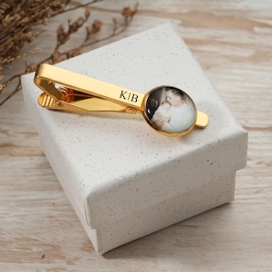 May include: A gold-colored tie clip with a circular photo and the initials "KIB". The photo shows a person holding a baby. The tie clip is resting on a white gift box.