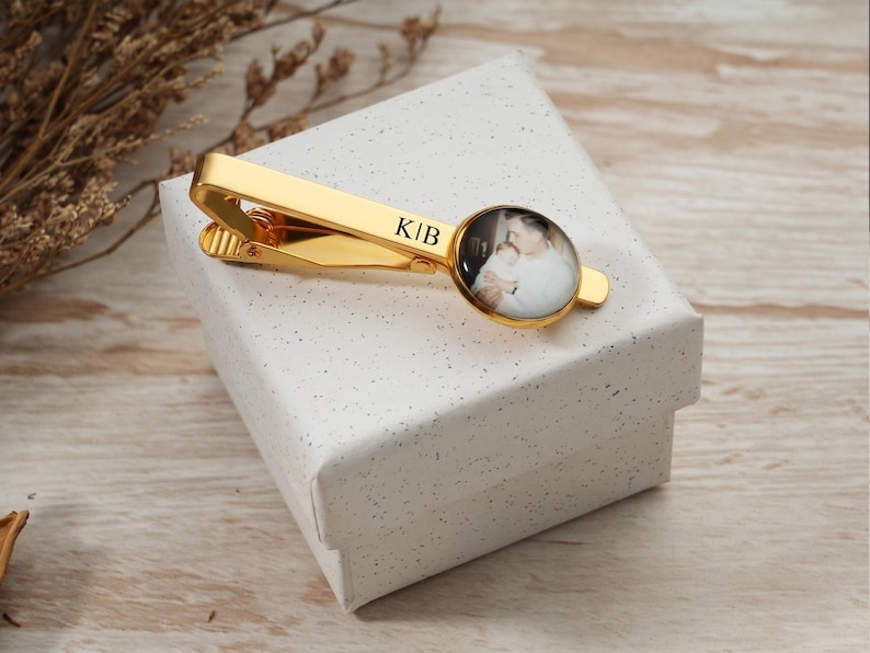 May include: A gold-colored tie clip with a circular photo and the initials "KIB". The photo shows a person holding a baby. The tie clip is resting on a white gift box.