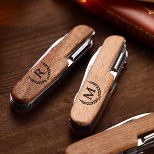 Puede incluir: Primer plano de navajas de bolsillo de madera personalizadas. Las navajas tienen un mango de madera marr&oacute;n claro con una hoja y herramientas plateadas. Cada navaja est&aacute; grabada con una letra dentro de una corona de laurel. El fondo es una superficie de madera oscura.
