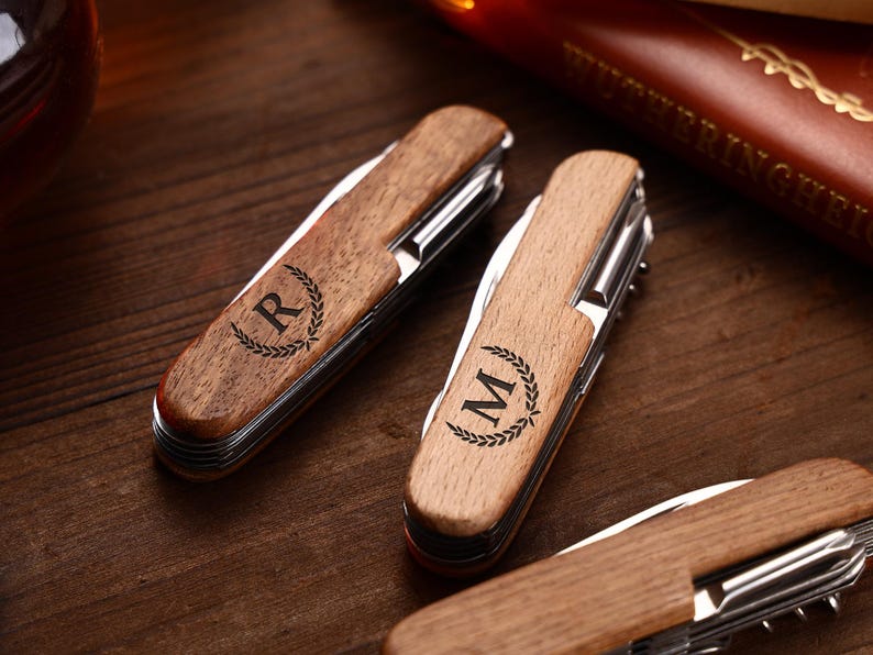 Puede incluir: Primer plano de navajas de bolsillo de madera personalizadas. Las navajas tienen un mango de madera marr&oacute;n claro con una hoja y herramientas plateadas. Cada navaja est&aacute; grabada con una letra dentro de una corona de laurel. El fondo es una superficie de madera oscura.