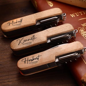 Puede incluir: Tres multiherramientas de madera con grabado personalizado. Cada herramienta tiene un mango de madera clara e incluye un cuchillo, un destornillador y otras herramientas. Los nombres "Michell", "Kenneth" y "Herbert" est&aacute;n grabados en los mangos, junto con el texto "Groomsman" y una fecha.