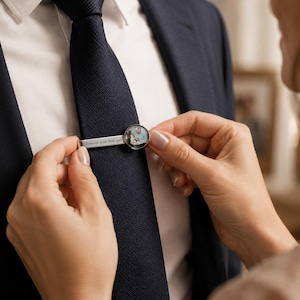 May include: A silver tie clip with a circular design and the text "Forever your little girl." The tie clip is being placed on a navy blue tie. The circular design appears to contain a small photograph.