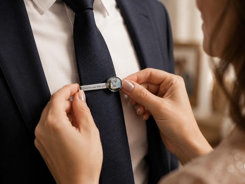 May include: A silver tie clip with a circular design and the text "Forever your little girl." The tie clip is being placed on a navy blue tie. The circular design appears to contain a small photograph.