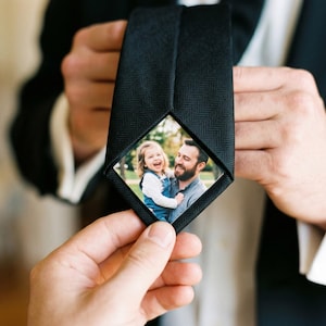 May include: Black tie with a diamond-shaped photo insert. The photo shows a man holding a young child. The tie has a textured pattern and is held by a person wearing a suit.