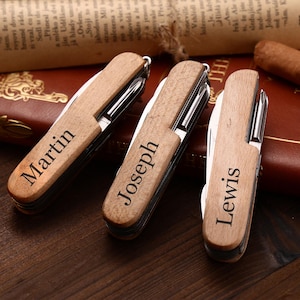 Puede incluir: Tres navajas de bolsillo de madera personalizadas, cada una grabada con un nombre: "Martin", "Joseph" y "Lewis". Las navajas est&aacute;n cerradas, mostrando el mango de madera. El fondo incluye un libro encuadernado en cuero y un cigarro.