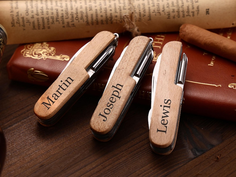 Puede incluir: Tres navajas de bolsillo de madera personalizadas, cada una grabada con un nombre: "Martin", "Joseph" y "Lewis". Las navajas est&aacute;n cerradas, mostrando el mango de madera. El fondo incluye un libro encuadernado en cuero y un cigarro.