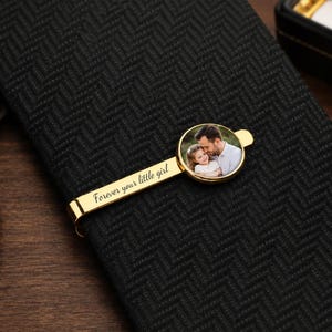 May include: A gold-colored tie clip with a circular photo of a man and a child. The clip is on a black herringbone-patterned tie. The text "Forever your little girl" is engraved on the clip.