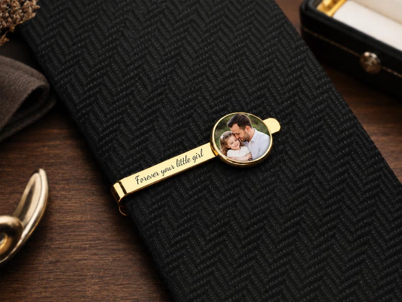 May include: A gold-colored tie clip with a circular photo of a man and a child. The clip is on a black herringbone-patterned tie. The text "Forever your little girl" is engraved on the clip.