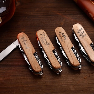Puede incluir: Cuatro navajas de bolsillo de madera con grabados personalizados, incluyendo nombres e iniciales. Cada navaja presenta un sacacorchos y una hoja plateados, sobre una superficie de madera oscura. Una botella de licor es visible en el fondo.