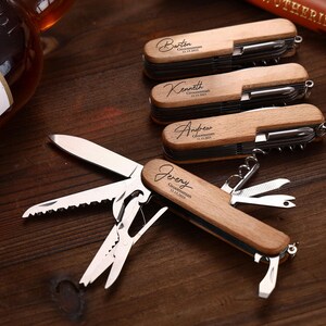 Puede incluir: Multiherramientas de madera con grabado personalizado. Cada herramienta cuenta con cuchillo, sierra, tijeras y otros implementos. Los nombres "Barton", "Kenneth", "Andrew" y "Jeremy" est&aacute;n grabados en las empu&ntilde;aduras de madera, junto con "Groomsman" y una fecha.