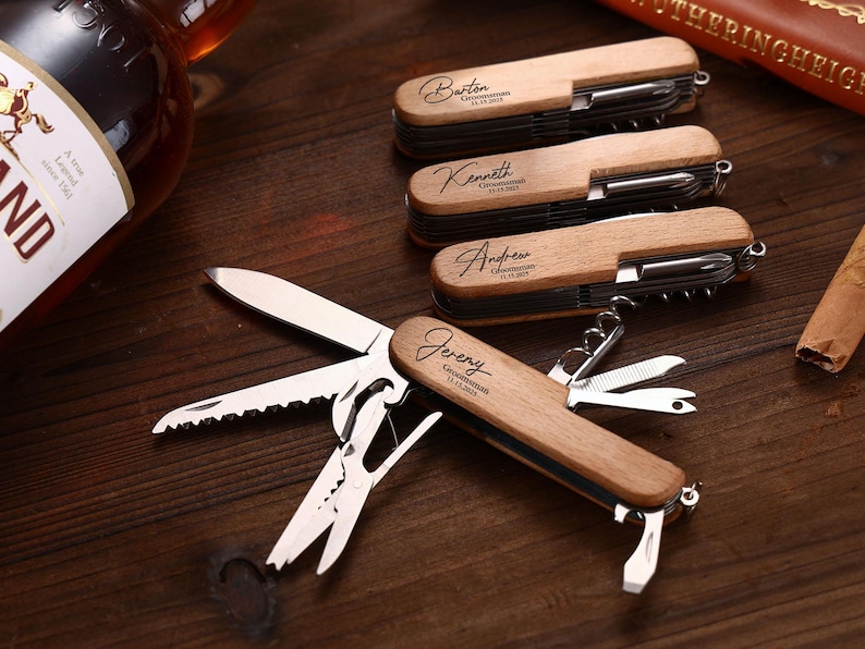 Puede incluir: Multiherramientas de madera con grabado personalizado. Cada herramienta cuenta con cuchillo, sierra, tijeras y otros implementos. Los nombres "Barton", "Kenneth", "Andrew" y "Jeremy" est&aacute;n grabados en las empu&ntilde;aduras de madera, junto con "Groomsman" y una fecha.