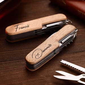 Puede incluir: Dos multiherramientas de madera con componentes de acero inoxidable. Una est&aacute; grabada con "Francis" y la otra con "Dannie". Las herramientas est&aacute;n sobre una superficie de madera oscura, con herramientas adicionales visibles.