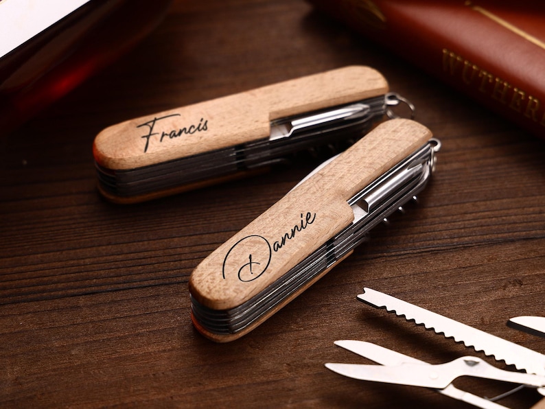 Puede incluir: Dos multiherramientas de madera con componentes de acero inoxidable. Una est&aacute; grabada con "Francis" y la otra con "Dannie". Las herramientas est&aacute;n sobre una superficie de madera oscura, con herramientas adicionales visibles.