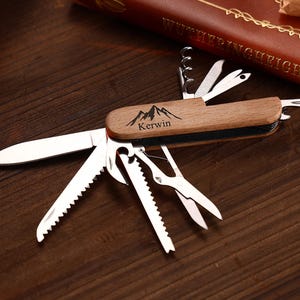 Puede incluir: Una multiherramienta con mango de madera y varios implementos de acero inoxidable, incluyendo un cuchillo, sierra, tijeras y sacacorchos. El mango est&aacute; grabado con un dise&ntilde;o de monta&ntilde;a y el nombre "Kerwin".