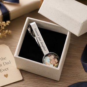 May include: A silver tie clip with the words "Forever your daughter" engraved on it, presented in a gift box. The clip features a photo of a father and daughter. A gift tag reads "For the Best Dad Ever."