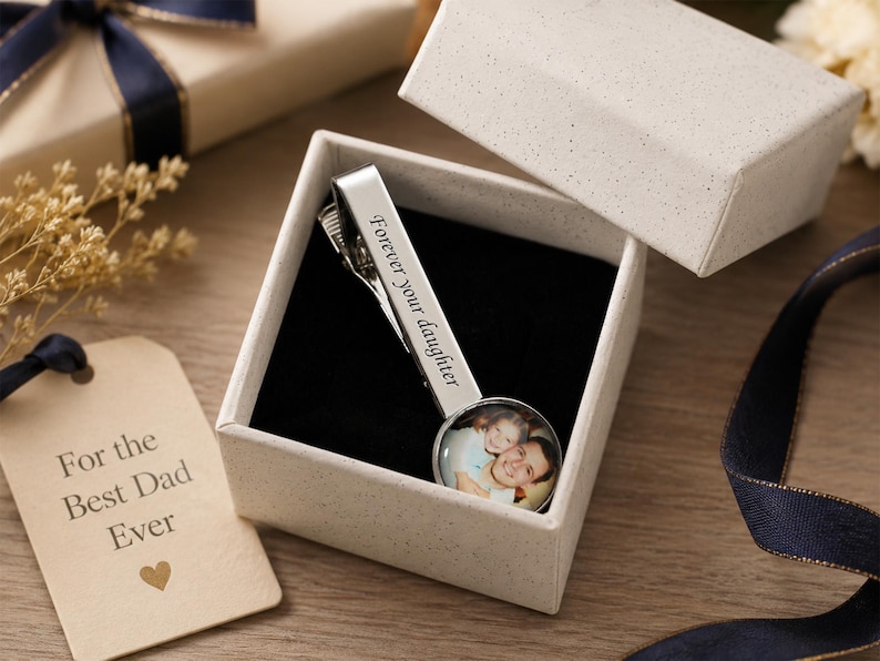 May include: A silver tie clip with the words "Forever your daughter" engraved on it, presented in a gift box. The clip features a photo of a father and daughter. A gift tag reads "For the Best Dad Ever."