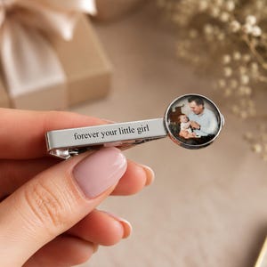 May include: Silver tie clip with the inscription "forever your little girl." The clip features a circular image of a man holding a baby. The background includes a gift box and floral accents.