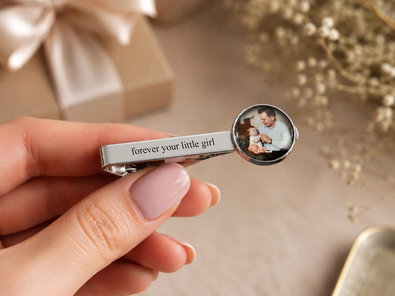 May include: Silver tie clip with the inscription "forever your little girl." The clip features a circular image of a man holding a baby. The background includes a gift box and floral accents.