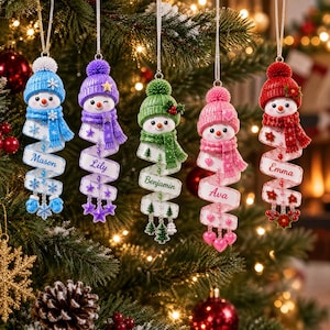 May include: Five hanging snowman ornaments with knitted hats and scarves in blue, purple, green, pink, and red. Each snowman features a spiral ribbon body personalized with the names Mason, Lily, Benjamin, Ava, and Emma, accented with themed charms.
