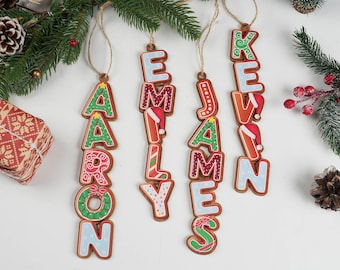 May include: Gingerbread-style Christmas ornaments spelling out names: Aaron, Emily, James, and Kevin. Each letter is decorated with festive colors and patterns, some with Santa hats. The ornaments hang from twine.