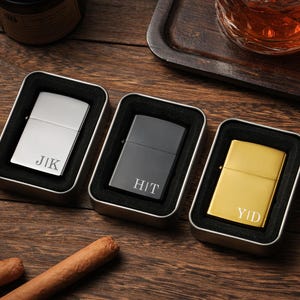 May include: Three personalized lighters in metal tins, each with initials engraved. One is silver, one is black, and one is gold. The lighters are displayed on a wooden surface, with cigars and a glass of amber liquid nearby.